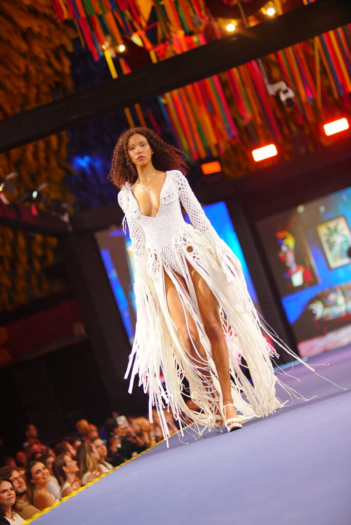 Modelo caminha na passarela usando um vestido branco de macramê com mangas longas, decote profundo e uma saia composta por longas franjas de fios que vão até os pés. Ao fundo, bandeirinhas coloridas decoram o teto e o público observa dos dois lados da passarela.