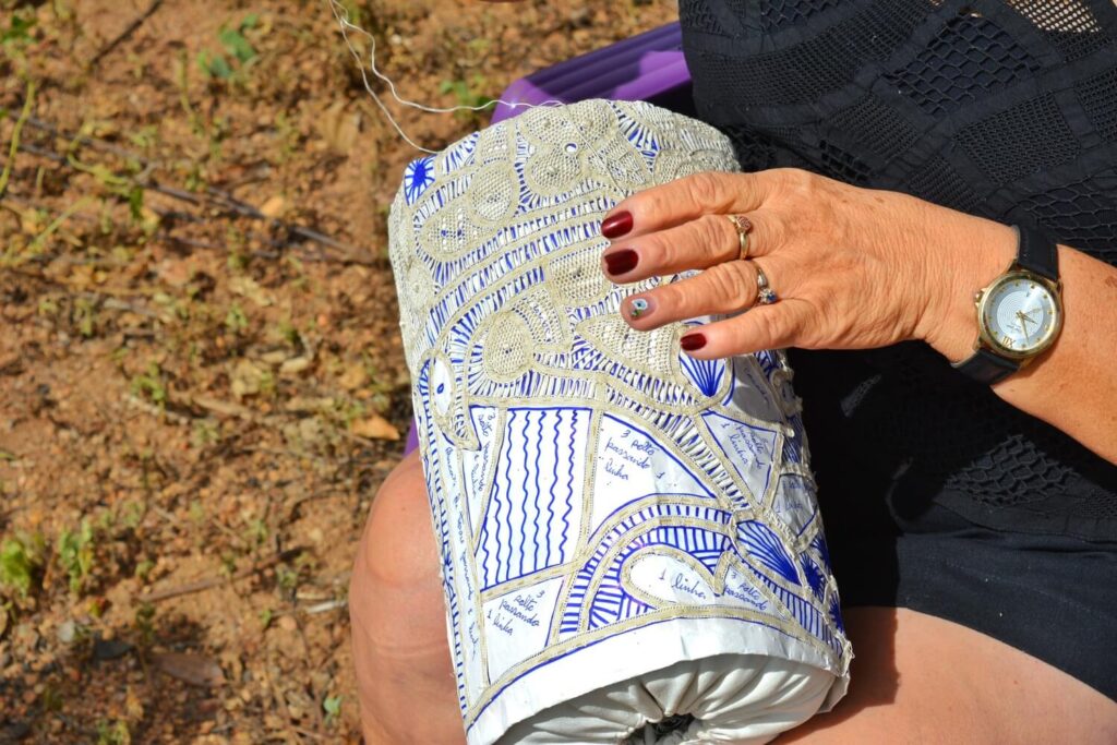 Uma pessoa sentada ao ar livre segura sobre o colo uma almofada cilíndrica usada na confecção da renda renascença. Sobre o molde de papel com desenhos geométricos, fios claros são entrelaçados manualmente, formando a renda. 
