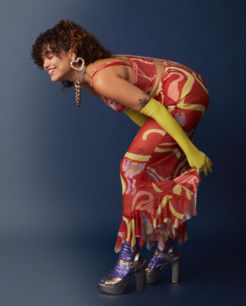 Modelo negra de cabelo cacheado está curvada sorrindo no fundo azul petróleo usando vestido levemente transparente e colorido com as cores rosa e verde limão, e também usa luvas até o cotovelo da cor verde limão.