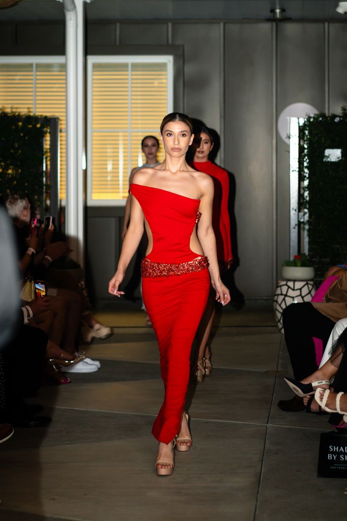 Model in a striking red dress walks a fashion runway, showcasing elegance and style.