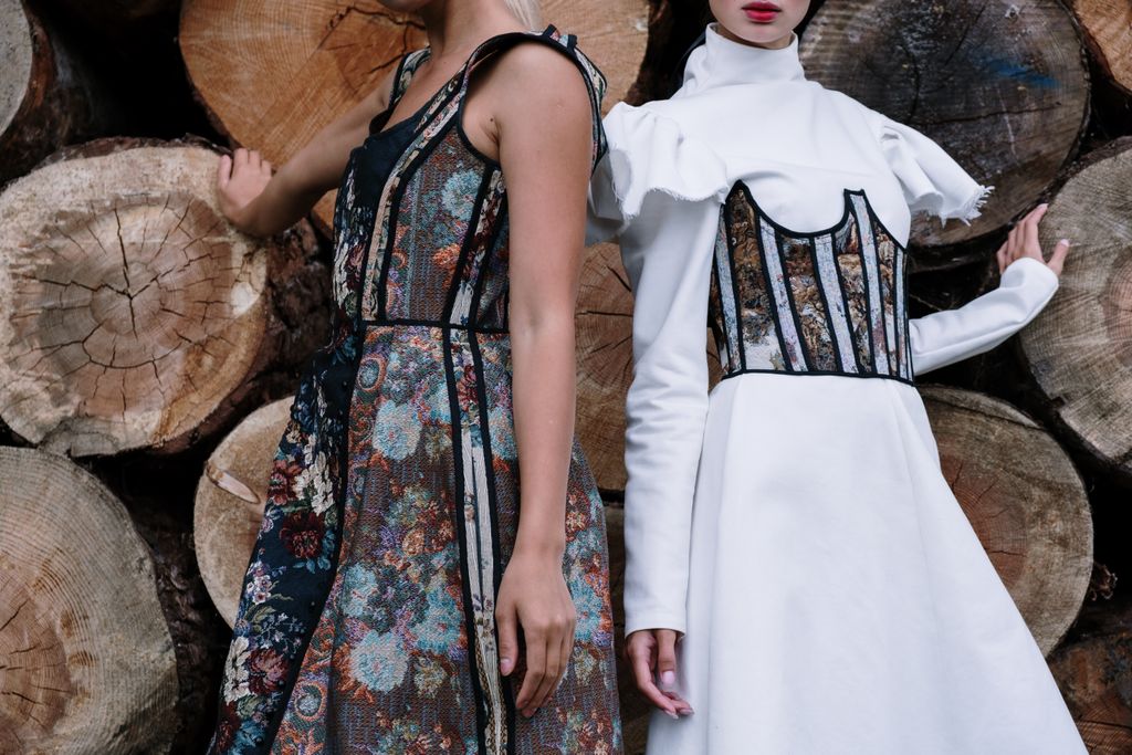 Two women in floral dresses pose in front of stacked wood logs, showcasing intricate couture fashion.